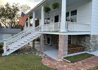 White house with a front porch, American flag, brick steps, and hanging plants, surrounded by grass and trees.