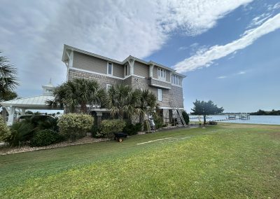 A large beach house with palm trees in front, set beside a grassy lawn and waterfront under a partly cloudy sky.