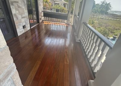 A polished wooden porch with white railings overlooks a yard and distant houses on a sunny day.