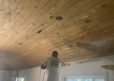 Person painting a wooden ceiling with a roller in a sunlit room, ladder and windows visible.