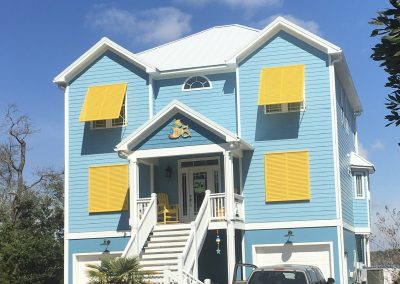 A blue house with yellow shutters, white trim, and a front porch, with a car parked in the driveway.