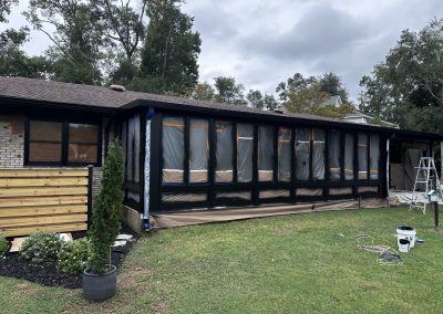 A house sunroom is covered in plastic and painter's tape, with painting supplies on the lawn nearby.