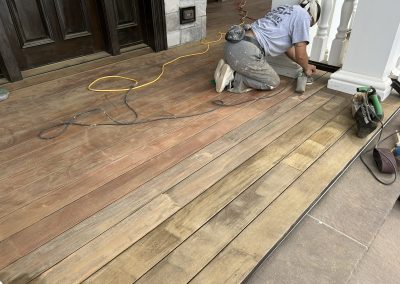 A person sanding and refinishing a wooden porch floor near a white railing and brown door.