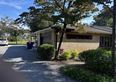 A brick house with a carport, trees, and shrubs along a gravel driveway on a sunny day.