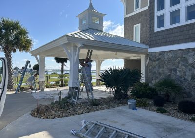 A person on a ladder paints a white gazebo attached to a house, with palm trees and water in the background.