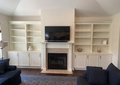 White built-in shelves and cabinets flank a fireplace with a TV above, in a living room with blue sofas.