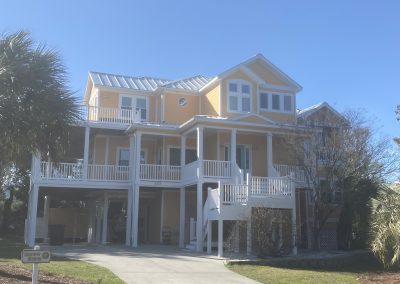 A large yellow beach house with white trim, balconies, and a driveway surrounded by palm trees.