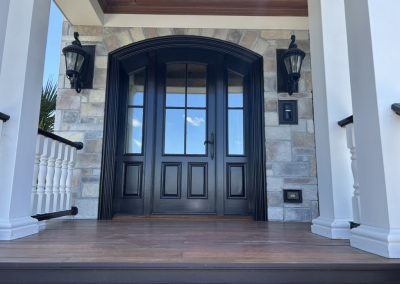 Elegant black front door with glass panels, framed by stone wall and two black wall lanterns on a porch.