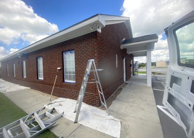 A brick building being painted with ladders and drop cloths set up outside under a partly cloudy sky.