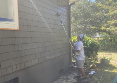 Person painting the side of a house with a roller on a long pole on a sunny day.