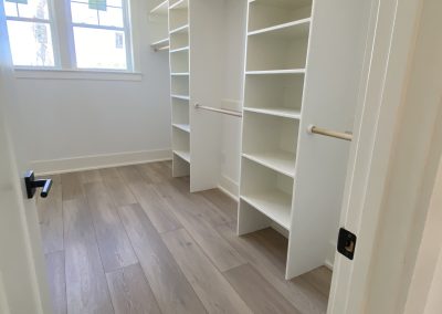 Empty walk-in closet with wooden floor, white shelves, and hanging rods, lit by two windows.