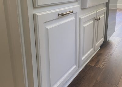White kitchen cabinets with silver handles and a farmhouse sink, set on wood flooring.