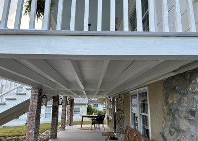 Covered patio with stone wall, bench, lanterns, and outdoor table, beneath a white wooden balcony.