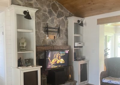 Living room with stone fireplace, TV, built-in shelves, wood ceiling, and wicker chair on a patterned rug.