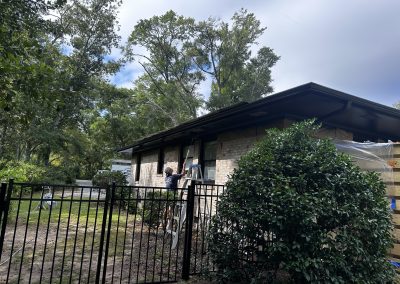 Person on a ladder painting the exterior of a light brick house, surrounded by trees and a black fence.