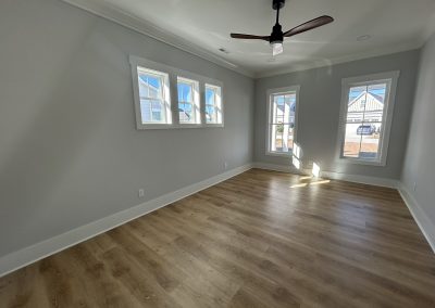 Empty, sunlit room with light wood flooring, gray walls, three windows, and a ceiling fan.