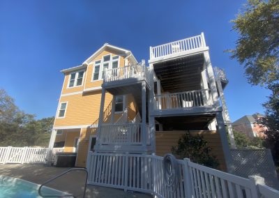 Three-story yellow house with white trim and balconies, next to a white fence and pool under a clear blue sky.