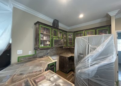 Kitchen under renovation with cabinets taped and covered, appliances wrapped in plastic, and paint supplies visible.