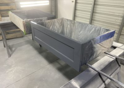 Gray drawer boxes covered in plastic are being spray painted in a workshop with metal walls.