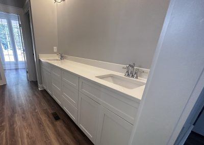 Long white bathroom vanity with two sinks, chrome faucets, and white cabinets, next to a wood floor hallway.