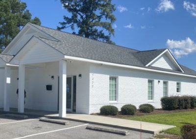 White one-story building with columns, surrounded by shrubs and trees under a blue sky with clouds.