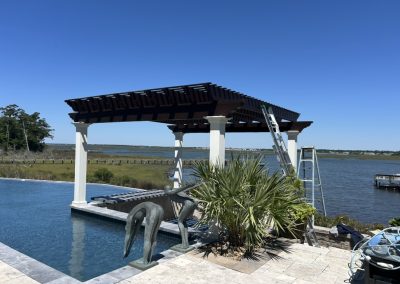 A pergola by a pool overlooks a lake; a statue, ladder, and plants are nearby under a clear blue sky.