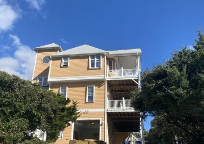 Three-story tan house with white trim, viewed from the driveway, surrounded by trees under a blue sky.