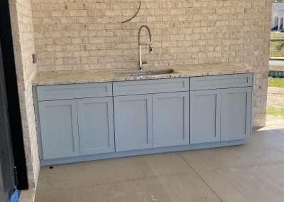 Light blue outdoor kitchen cabinets with a marble countertop, sink, and a brick wall background.