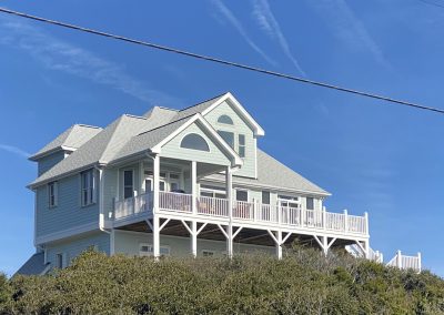 Light blue two-story house with a wraparound porch on a hill, under a clear blue sky with wispy clouds.
