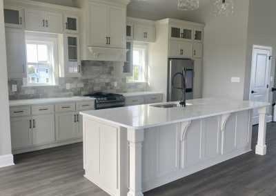 Modern white kitchen with island, stainless steel appliances, and pendant lights over gray wood flooring.