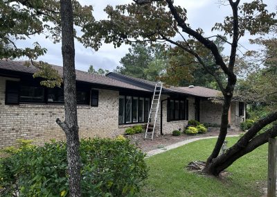 A ladder leans against the roof of a brick house surrounded by trees and green shrubs.