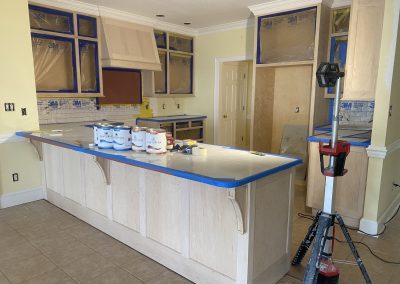 Kitchen under renovation with cabinets taped off, paint cans on island, and protective coverings in place.