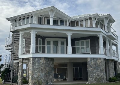 Three-story beach house on stone pillars with wraparound balconies and a spiral staircase outside.