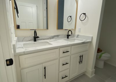 Modern bathroom with double sinks, arched mirrors, black fixtures, and light gray tile floor. Toilet visible at right.
