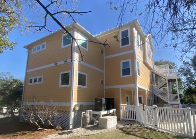A three-story yellow house with white trim, a fenced yard, and exterior staircases on a sunny day.