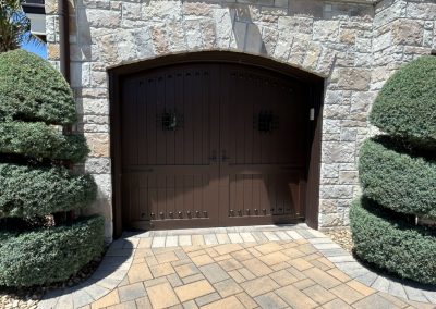 Large brown double doors with metal accents set in a stone wall, flanked by trimmed green bushes.