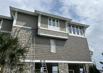 Two ladders lean against a raised beach house with gray siding under a partly cloudy sky.