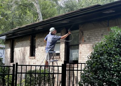 A person on a ladder paints the trim of a brick house with a spray gun near trees and a metal fence.