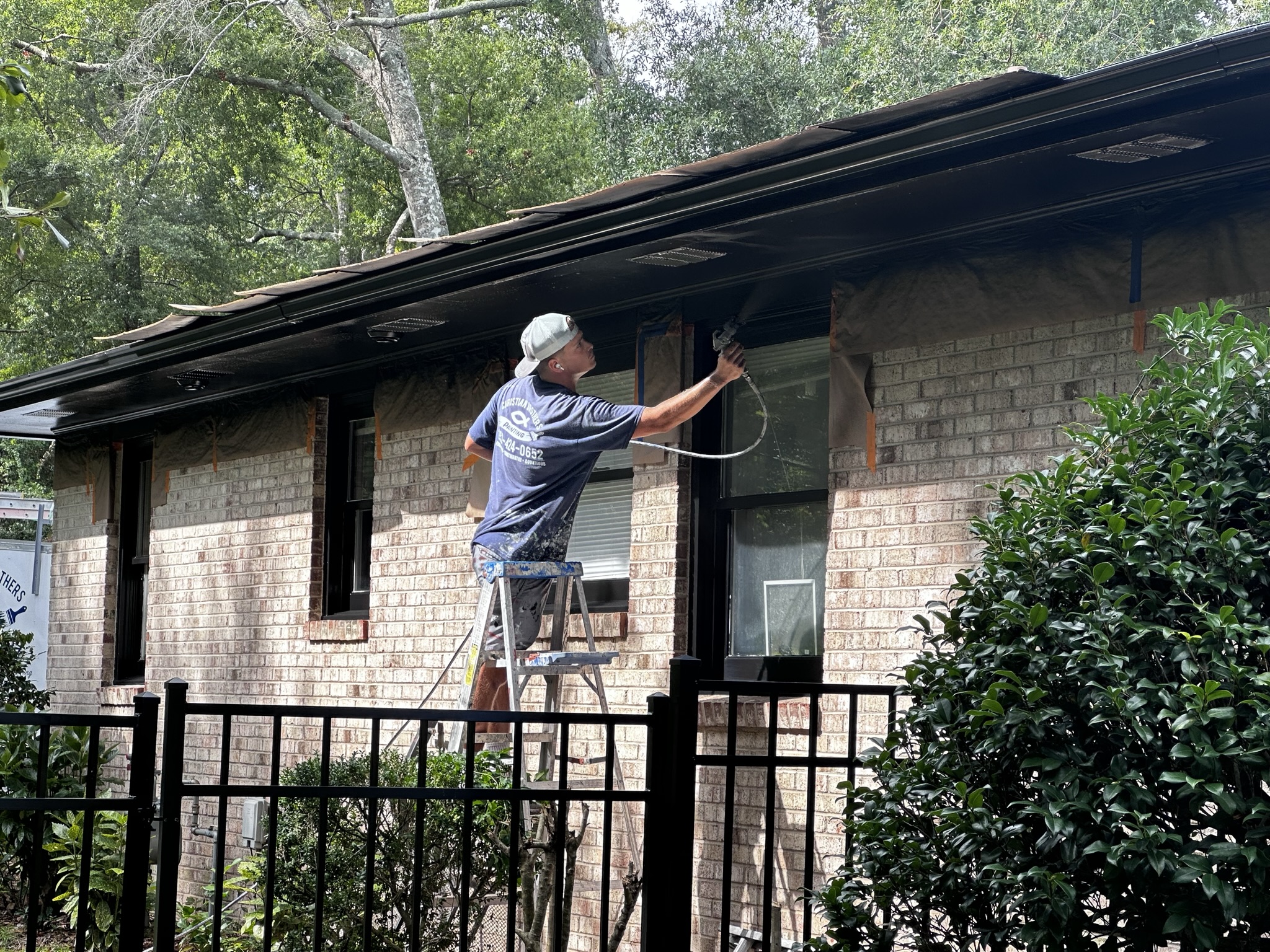 CC49E751-A585-4E7B-B2F6-D3E61F1CCE0C_1_102_o A person on a ladder paints the trim of a brick house with a spray gun near trees and a metal fence.