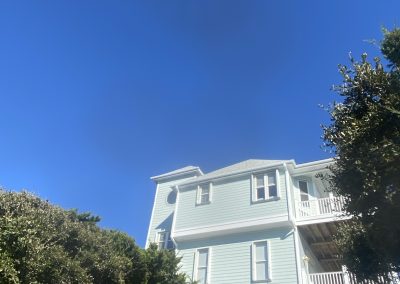 Light blue three-story house with balconies, surrounded by greenery, under a clear bright blue sky.