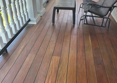 A porch with reddish wood flooring, a black mesh chair, and a small black table beside white railings.