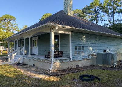 Light blue house with a covered porch, porch swing, and trees in the background under a clear sky.