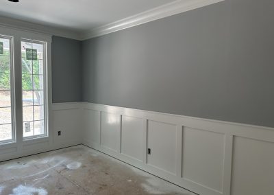 A room with gray walls, white wainscoting, large windows, and unfinished floors.