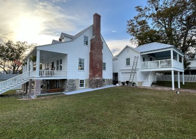 White farmhouse with a red brick chimney, porch, and attached building on a grassy lawn at sunset.