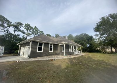 Single-story gray house with a porch and garage, surrounded by trees and a grassy yard under a cloudy sky.