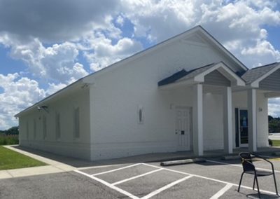 A small white brick building with a porch, under a partly cloudy sky, with an empty parking lot in front.