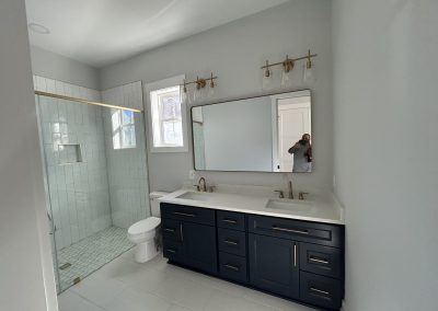 Modern bathroom with a glass shower, double vanity, and gold fixtures; person seen in mirror.