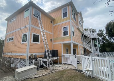 A tall ladder leans against a peach-colored house; paint supplies are on the ground under a cloudy sky.