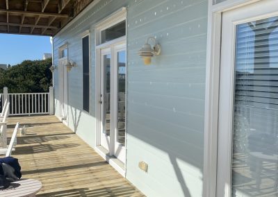 Sunny wooden deck with light blue siding, white trim, outdoor lights, and chairs; trees in the background.